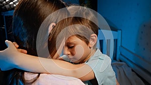 Portrait of little upset and sad toddler boy hugging and kissing his mother before going to sleep at night. Loving and