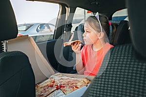 Portrait of little 5 YO girl eating just cooked Italian pizza sitting in child car seat on car back seat and looking out window .
