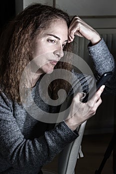 Portrait of lonely mature woman with worried facial expression thinking holding her mobile during quarantine lockdown