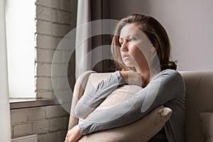 Portrait of lonely sad Ñaucasian young woman sitting near window and hugs pillow.