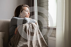Portrait of lonely sad Ñaucasian young woman wrapped in a plaid sitting near window of apartments.