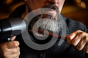 Portrait of a long bearded man with a hairdryer and a round comb at home, closeup