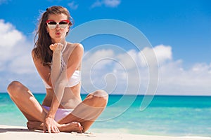 Portrait of long haired woman in white bikini and sunglasses blowing a kiss on tropical beach. La Digue, Seychelles