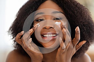 Portrait of lovely black woman applying face cream onto her cheeks, smiling at camera, indoors