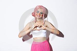 Portrait of lovely girl in pink party wig and bright makeup, showing heart gesture and smiling, standing over white