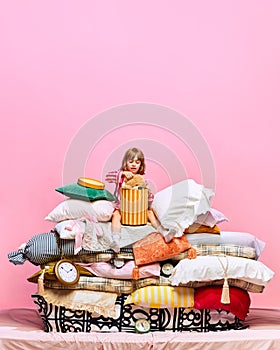 Portrait of lovely little princess unpacking present with teddy bear sitting feather bed with pillows over pink studio