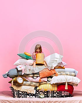 Portrait of lovely little princess unpacking presents sitting feather bed with pillows over pink studio background.