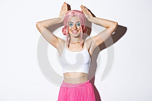 Portrait of lovely party girl in pink anime wig, showing bunny ears and smiling happy, celebrating halloween, standing