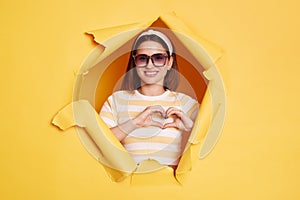 Portrait of lovely romantic woman wearing striped shirt and hair band, breaks through yellow paper background, showing heart shape