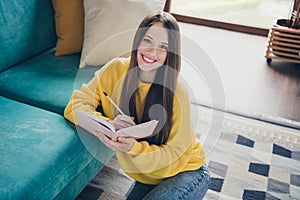 Portrait of lovely satisfied young girl toothy smile sitting carpet floor hold pencil notepad bright apartment inside