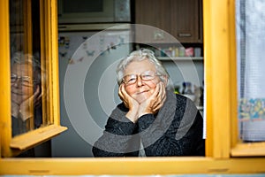 Portrait of lovely senior woman or grandmother watching out from the window and smiling, pensioner