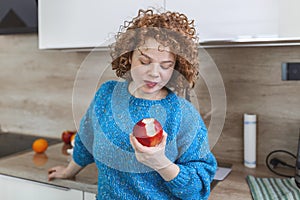 Portrait of a lovely smiling pretty girl biting an apple in her kitchen. Young woman enjoying her red organic apple fruit. Daily