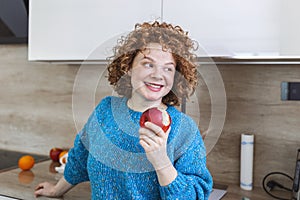 Portrait of a lovely smiling pretty girl biting an apple in her kitchen. Young woman enjoying her red organic apple fruit. Daily