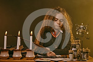 Little witch dressed in dark clothes sitting at the table against a black smoky background. There are magic wand, books