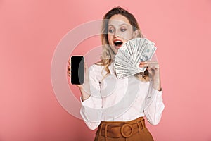 Portrait of lovely woman holding cell phone and money fan isolated over pink background