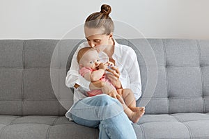 Portrait of lovely young adult woman with bun hairstyle wearing white shirt and jeans, sitting on sofa with baby drinking water