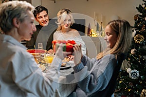 Portrait of loving grandmother giving festive box with Christmas present to adorable granddaughter sitting at dinner