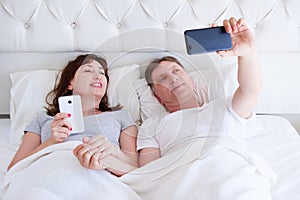 Portrait of loving middle aged couple smiling and make selfie on smartphone in bedroom, happy family