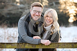 Portrait Of Loving Newly Engaged Couple Leaning On Fence On Snowy Walk In Winter Countryside