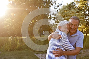 Portrait Of Loving Senior Couple Hugging Outdoors In Summer Park Against Flaring Sun