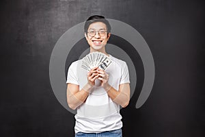 Portrait of lucky satisfied young rich asian male student holding lots of cash, fan of dollars smiling happy and pleased