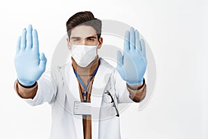 Portrait of mal doctor in face mask and robe, extend hands in gloves, showing stop gesture, taboo disapproval sign