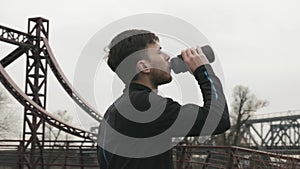 Portrait of male athlete drinking water from bottle. Caucasian young man in black sports clothes drinking energy drink from bottle