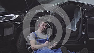 Portrait of male Caucasian auto mechanic in headphones rubbing eyes leaning against car wheel and using smartphone