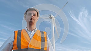 Portrait: the male engineer projects the work near the wind turbine. Sunny day and clouds. The man is dressed in a red