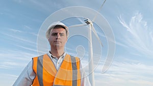 Portrait: the male engineer projects the work near the wind turbine. Sunny day and clouds. The man is dressed in a red