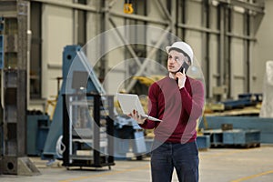 Portrait of a male factory manager in a white hard hat and red sweater holding laptop and mobile phone.