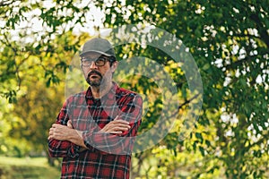 Portrait of male farmer with arms crossed wearing plaid shirt and trucker`s hat posing in walnut orchard and looking at camera