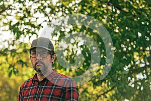 Portrait of male farmer wearing plaid shirt and trucker`s hat posing in walnut orchard and looking at camera