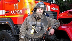 Portrait of male firefighter in helmet and balaclava standing near a big red car. Young fireman in full equipment