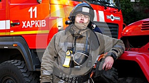 Portrait of male firefighter in helmet and balaclava standing near a big red car. Young fireman in full equipment