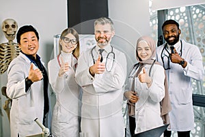 Portrait of male mature doctor with multiethnic medical team, smiling and showing thumbs up, against modern lab and