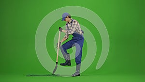Portrait of male in overalls and rubber boots on chroma key green screen. Man gardener digging a hole in the ground with