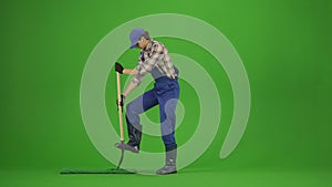 Portrait of male in overalls and rubber boots on chroma key green screen. Man gardener digging a hole in the soil with