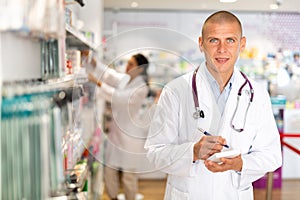 Portrait of male pharmacist working in pharmacy during the pandemic, standing in trading floor and makes important notes