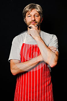 Portrait of a male professional butcher on a dark background. The model is in his 40s, grey hair and beard, dressed in red and