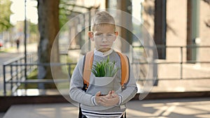 Portrait of the male pupil standing near school carying backpack wearing glasses holding the plant an a pot. People