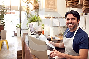 Portrait Of Male Sales Assistant Working On Laptop Behind Sales Desk Of Florists Store