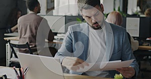 Portrait of male specialist reading business documents and working with laptop in open space office