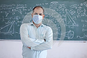 Portrait of male teacher wearing mask standing against blackboard teaching mathematics in classroom, school reopen after lockdown