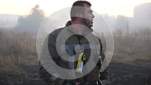 Portrait of male tired firefighter in uniform at smoky lawn after fire. Young fireman standing at countryside against