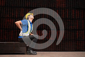 Portrait of a male worker wearing a safety vest and helmet sitting on a steels pallet due to back pain