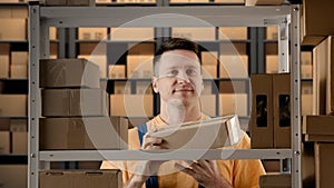 Portrait of male working in storage. Man storekeeper standing near rack with boxes, holding package in hand shows thumbs