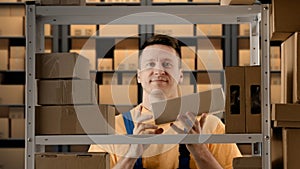 Portrait of male working in storage. Man storekeeper standing near rack with boxes, holding package in hand shows thumbs