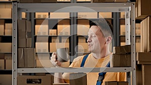 Portrait of male working in storage. Man storekeeper standing near rack taking mug drinks coffee enjoys, talking smiling