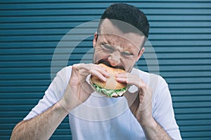 Portrait of man biting a piece of hamburger. He is shrinking. Isolated on striped and blue background.